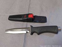 Tekna diver's cutter knife with black and red sheath lying on a flat surface, showing blade and handle details.