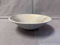 Side view of the shallow pottery bowl showing its flared rim and matte light neutral glaze with faint brown and black abstract linear designs.