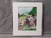 Framed watercolor depicting a village scene including houses with balconies, a street lamp, trees, hills, and a bridge over water in the distance.