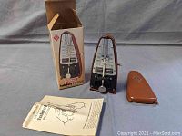 Wittner Taktell Piccolo metronome displayed with its original box, plastic cover, and instruction booklet on a grey background.