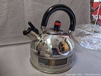 Photo showing the shiny stainless steel kettle with black curved handle and spout lever. Red accent on lid knob, kettle marked 'Profes'.