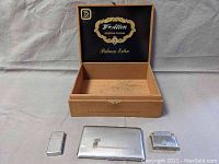 Wooden cigar box open showing interior branding with three items placed in front: silver cigarette case and two silver-tone lighters.