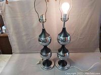 Two Mid-Century Modern chrome and black table lamps side by side, showing entire fixture from base to light socket, one lamp lit.