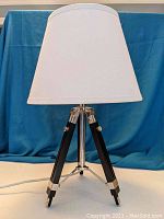 Clear image showing the tripod base of the lamp with three wooden legs and metal accents, turned off with the shade white.