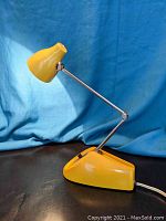 Yellow mid-century desk lamp with extendable arm and compact base on a black surface and blue background.