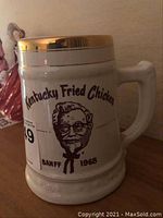 Side view of white ceramic KFC tankard with gold trim and printed graphics including Colonel Sanders portrait and Banff 1968 text.