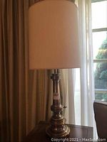 Front and side view of brass table lamp with lampshade, placed on wooden surface near window and curtains.