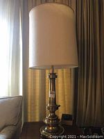 Full view of the vintage brass table lamp standing on a side table near a couch with natural light from a window behind it.