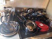 Overview of collection showing pots, pans, teapot, French press, mixing bowls, measuring cups, and red waffle maker on kitchen stove and counter.