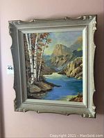 Front view of framed landscape painting featuring a river with birch trees, mountains in the background.