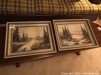 Two framed landscape oil paintings side by side on a bench