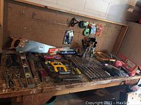 Overview of large assortment of hand tools laid on wooden table including saws, clamps, wrenches, pliers, screwdrivers and measuring tools with pegboard background.