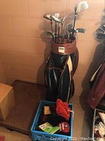 Golf bag filled with various golf clubs placed on the floor with a blue container of golf accessories nearby.