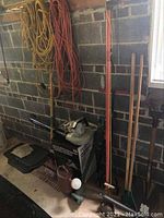 Photo of garden tools leaning against wall near window and concrete blocks. Shows coiled extension cords, long handled rakes, shovels, and a garden sprayer on floor.