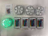 Four submersible RGB LED lights in clear packaging with four remote controls and instruction manual, one light illuminated green.