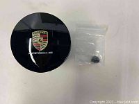 Closed black Porsche car ashtray showing the Porsche crest on the lid and a small accessory part in plastic bag beside it.