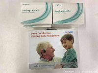 Four Sengdong Hearing Amplifiers sealed in box and Bonein Bone Conduction Hearing Aids Headphone box with image of elderly man and child