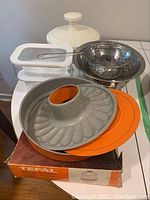 Photo showing T-Fal cake mold with orange silicone base, stainless steel strainers, white salad spinner, and white storage containers on a table.