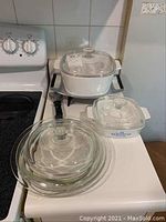 Photo showing two Corning Ware casserole dishes on stove: large white casserole with glass lid and metal handles, smaller blue floral patterned casserole with glass lid, and four clear glass Pyrex bowls.