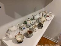 Table with multiple assorted tea pots, tea cups, and creamers arranged in a row showing different designs and sizes