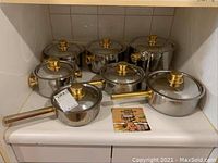 Photo showing full set of seven stainless steel pots with brass handles and lids arranged on counter with product brochure included