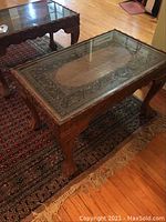 Front angled view showing full table, glass top, detailed carvings, claw feet, and rug beneath.