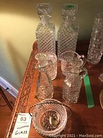 Full view of glassware set on wooden table, showing two large square decanters with stoppers, two smaller pitchers, and other pieces.