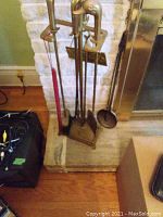 Photo of brass fireplace tools in a stand placed near a stone fireplace hearth showing the duck head handles and various tools.