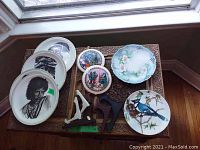 Overview of multiple decorative plates with artistic designs including a male portrait, floral, and bird illustrations along with several plate stands.