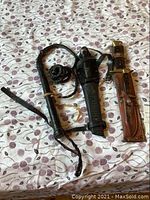 Photo showing a braided leather bull whip with loop handle, next to two cutters in leather sheaths, all laid on patterned fabric surface.