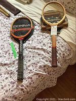 Two wooden tennis rackets laid on a floral bedspread, one Donnay with black grip and orange/black frame, the other Dunlop Maxply Fort with brown grip, wooden press in the frame, and black cover beneath.