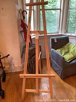 Front view of the wood folding easel standing upright on a hardwood floor near a window and armchair.