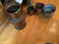 Photo showing five metalware pieces on wooden floor: a large copper pitcher, two smaller copper containers, a small copper pot, and a scalloped tin or galvanized metal basin.