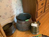 Three brass and metal ware items on a wooden floor next to a dresser: covered brass pitcher with handle, large round decorative metal container, smaller plain metal container.