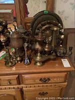 Photo showing a collection of various metal ware items on a wooden dresser including trays, oil lamp, pitchers, vases, and candle holders.