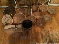 Photo showing assortment of 12 woven baskets varying in shape, size, and weave pattern with dark woven cylindrical basket in front and decorative wood duck box seen at bottom left.