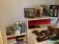 White bookcase with coffee table books on top and on the shelves, visible wear and tear, wooden carved decorations on side and top of the covered surface beside bookcase.