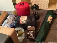 Red sleeping bag, black sleeping bag, green tent bag, black Coleman lantern, box with Feather 400 camping stove label, and other camping gear items visible.