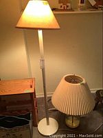 Photo showing both lamps: a tall floor lamp with metal base and beige shade, and a glass table lamp with pleated shade, placed on carpeted floor against wall.