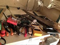 Photo showing assortment of hand tools including a large handsaw, clamp, hammers, and screwdrivers on a workbench or table.