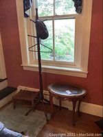 Wooden coat tree standing beside a window, small rush top stool partially visible next to it, and wooden side table with a glass top adjacent.