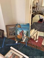 Original paintings and metal art stand viewed from a distance, focusing on overall lot contents, showing rolled paintings and rust on metal stand.