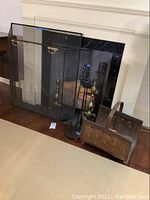 Fireplace screen in black mesh with brass handles, brass-like metal andirons, black fireplace tools on stand, and woven wood basket arranged before fireplace.