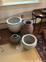 Four ceramic items including a large white planter with stand, two smaller round planters, and a tall pottery vase with floral design grouped on the floor next to a wall and table.