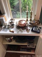 Full view of assorted metalware on a windowsill including copper stockpot, pewter and silver plate bowls, candlesticks, and small items like napkin rings and spoons.