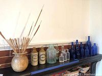 Blue and clear glass bottles, two stoneware jugs with labels, beige mottled vase with sticks on black shelf with brick wall background