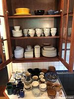 Full view of cabinet shelves and table with various white mugs, small plates, bowls, and other storage dishes mostly white with some blue patterns and brown oven dishes