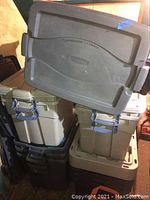 Photo showing a stack of multiple plastic storage totes in gray, black, and beige colors. The totes have lids with side locking clasps, some showing dirt and soiling from basement storage.