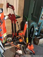 Photo showing multiple red electric weed whackers and hedge trimmers along with blowers leaned against wall in basement.