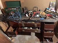 Workbench with drawers open showing tools inside, assorted hand tools on top including blue coiled cable, toolbox and vise visible.